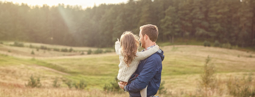 Concept image shows a father holding his daughter in a field bordered by a forest as the sun sets over trees in the background.